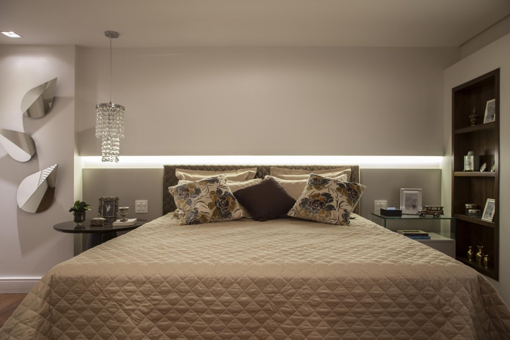 A suíte principal é dividida em feminino, com lustre de cristal e mesa de cabeceira redonda; e masculino com mesa em linhas retas e armário feito de imbuia. A iluminação com fita de led evidencia o recorte na parede de dry wall. Foto por J. Vilhora