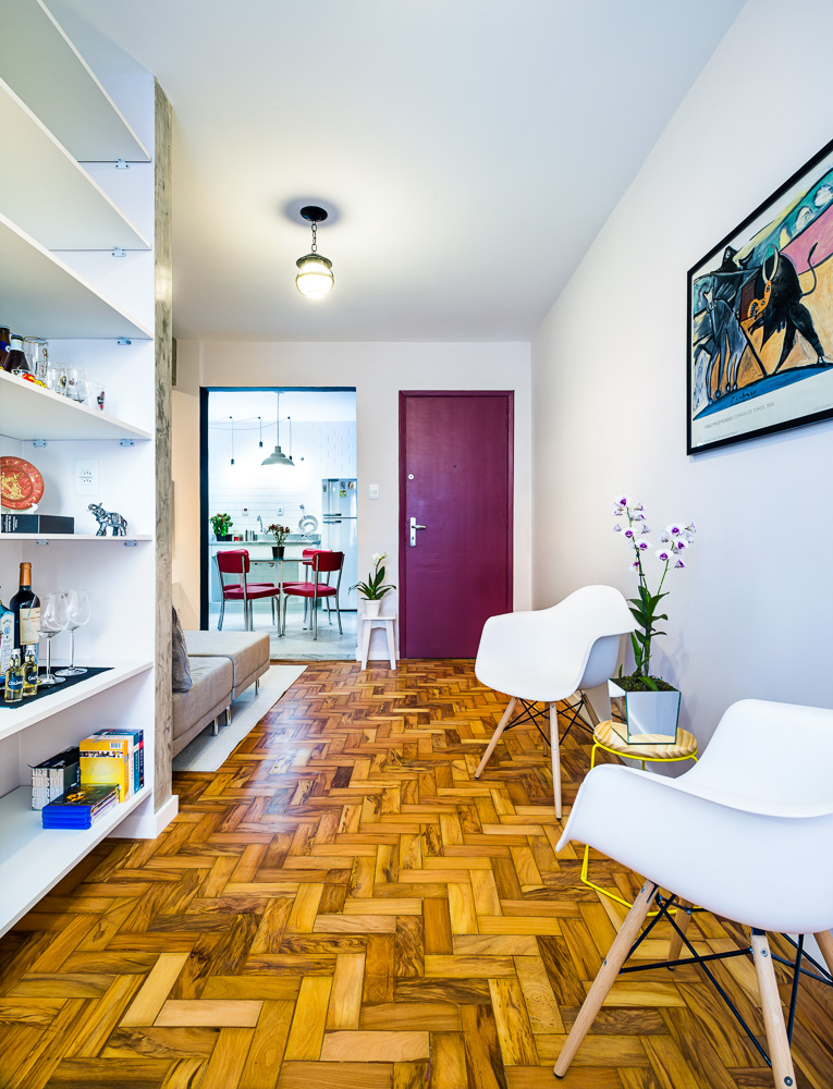 O piso original de madeira foi restaurado com a aplicação de nova resina, ganhando nova vida. Para compor a área do estar, duas cadeiras réplicas de Charles Eames (Oppa) trouxeram fluidez, tendo em vista as dimensões restritas do ambiente. Pinceladas de cores aparecem em alguns móveis e acabamentos, como a porta de entrada, que recebeu a cor Romã (Suvinil). Agora, a cozinha é integrada à sala graças à retirada da porta e à ampliação do vão. 