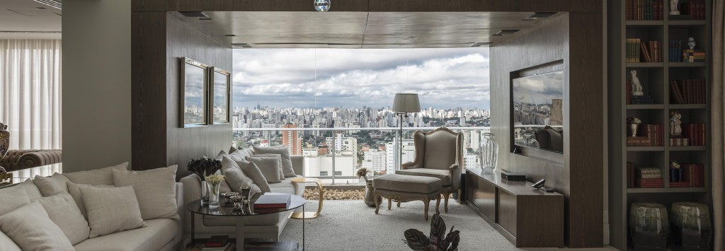 Na sala de estar com pé-direito duplo e vidros fixos do chão ao teto (Viplas), uma caixa de madeira escura (Marcenaria Barros) forma o ambiente de TV. O projeto de interiores, com sofás retilíneos, poltronas Luís XV e um grande lustre de cristal (Stylle), evidencia a mistura de estilos.