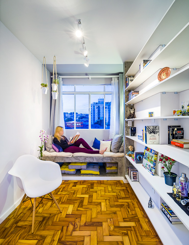 Para simular uma relaxante varanda, o projeto delimitou o espaço da janela com o banco de concreto coberto de futons (Oppa e Leroy Merlin) e destacou a área com cor diversa nas paredes e no teto. Para complementar, uma luminária tartaruga e pequenos vasos com espécies suculentas. Na parede contígua, uma estante desenhada sob medida auxiliou no aproveitamento de área. 