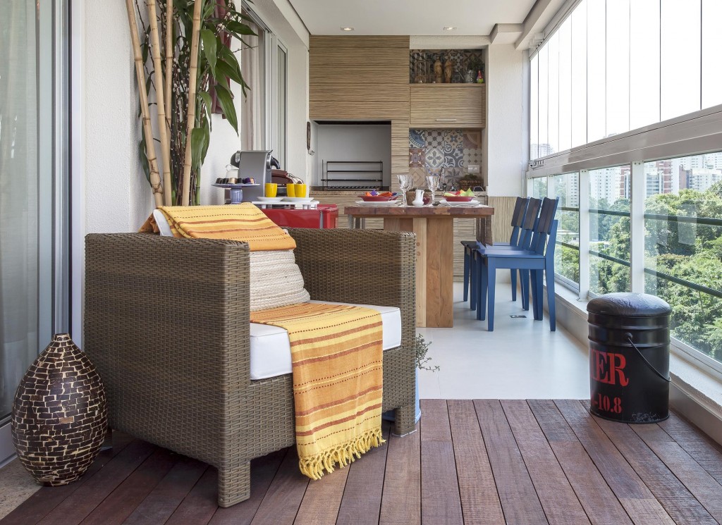 Na varanda a poltrona de fibra convida para se apreciar a vista voltada para a copa das árvores. Ao fundo junto da mesa, a marcenaria dá acabamento para a churrasqueira e a pia com frontão de porcelanato que reproduz ladrilho hidráulico.