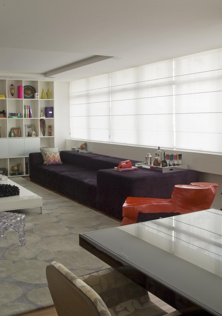 Do outro lado do estar, um jogo de cores: o tom berinjela no sofá de veludo harmoniza-se com o vermelho da mesa de tronco de árvore da designer Mônica Cintra. No piso, o tapete Samud em tom neutro. As cortinas romanas filtram a luz natural, abundante durante o dia, a área social é valorizada pela luz natural.