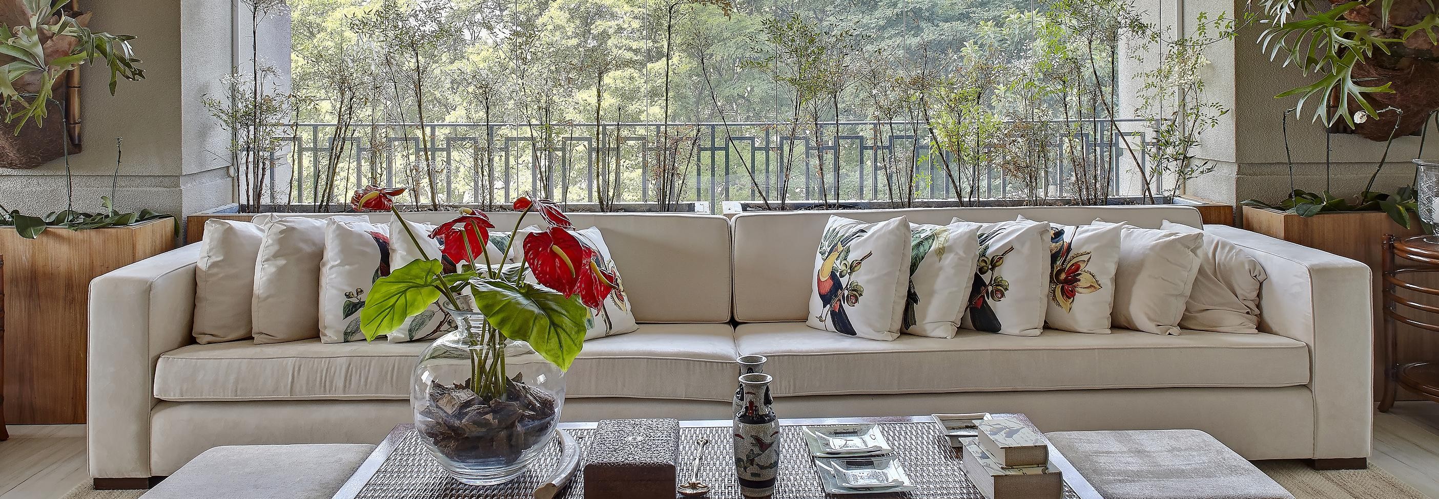 Detalhe da varanda fechada por vidro e conectada com o entorno verdejante. O farto sofá comporta muitas pessoas e é acompanhado pela mesa de fibras e dois pufes. Para enfatizar o estilo tropical, o paisagismo com espécies brasileiras e as almofadas com motivos de pássaros (JRJ Tecidos).