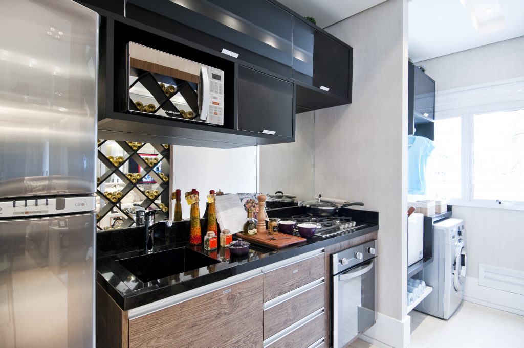 A cozinha é compacta e se conecta à lavandeira, ocupando toda a parede com área de trabalho, eletrodomésticos e armários planejados (Marel), bancada de granito preto absoluto, da Potenza Mármores.