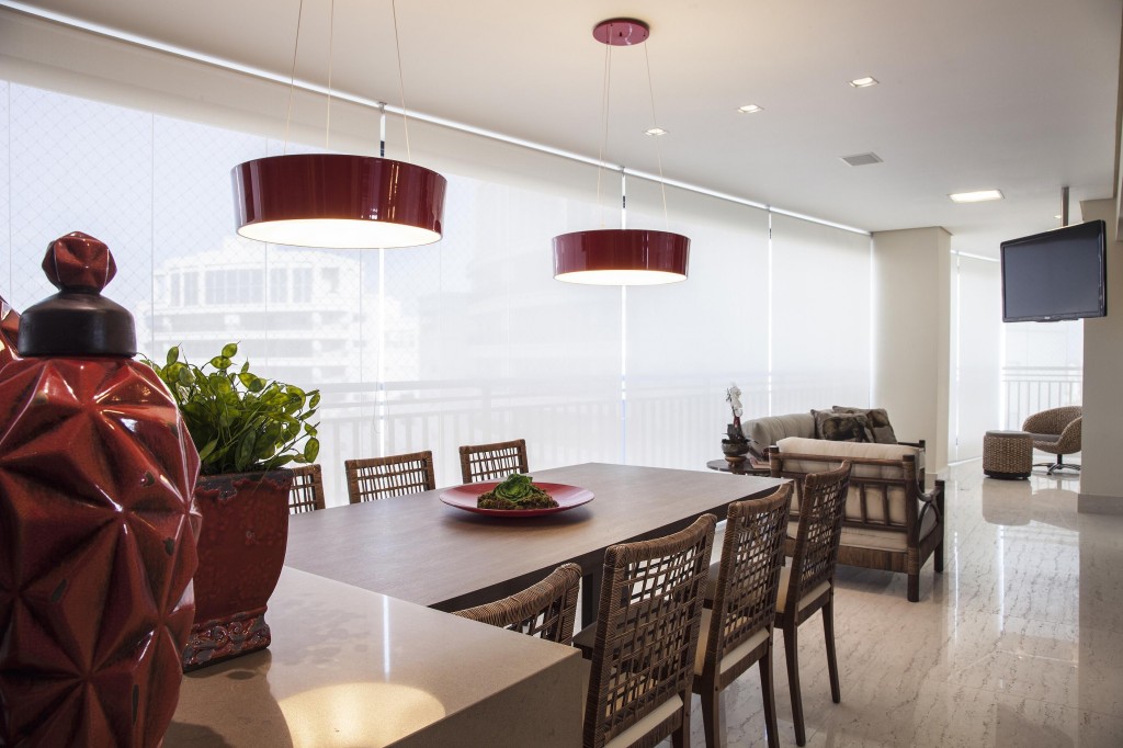 A área gourmet da varanda é definida pela bancada de onde sai a mesa de MDF padrão amadeirado feita sob medida que acomoda oito cadeiras com encosto de fibra. Cortinas em toda a extensão do vidro criam ambiente com iluminação suave. Foto por J. Vilhora