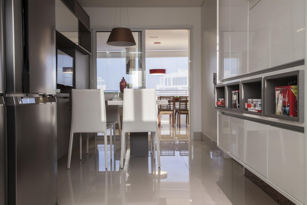 A cozinha diferencia-se pelo revestimento em porcelanato claro e homogêneo que contrasta ao modelo com padrão mármore. À esquerda, o móvel também acopla os eletrodomésticos. Já à direita, o armário suspenso acabado de laca branca brilhante contrasta com o cinza que também pincela os nichos abertos para deixar produtos sempre à mão. Foto por: J. Vilhora