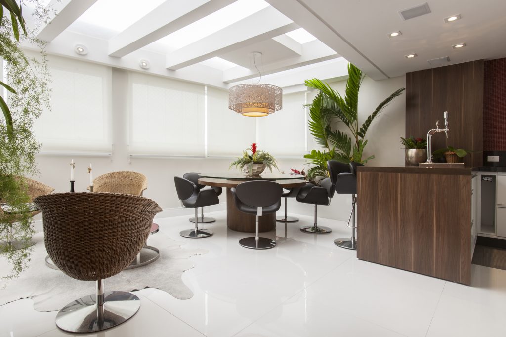 Ponto alto do imóvel com design modificado ainda na planta, a varanda é o lugar para receber os amigos. Com duas mesas de madeira e vidro acompanhadas de cadeiras de fibra, possui um pequeno bar com chopperia junto ao balcão que faz divisória com a cozinha. Os rasgos no teto deixam a luz entrar com abundância, multiplicada pelo piso branco. Nas janelas, as persianas (Beare Decor) controlam tamanha claridade.