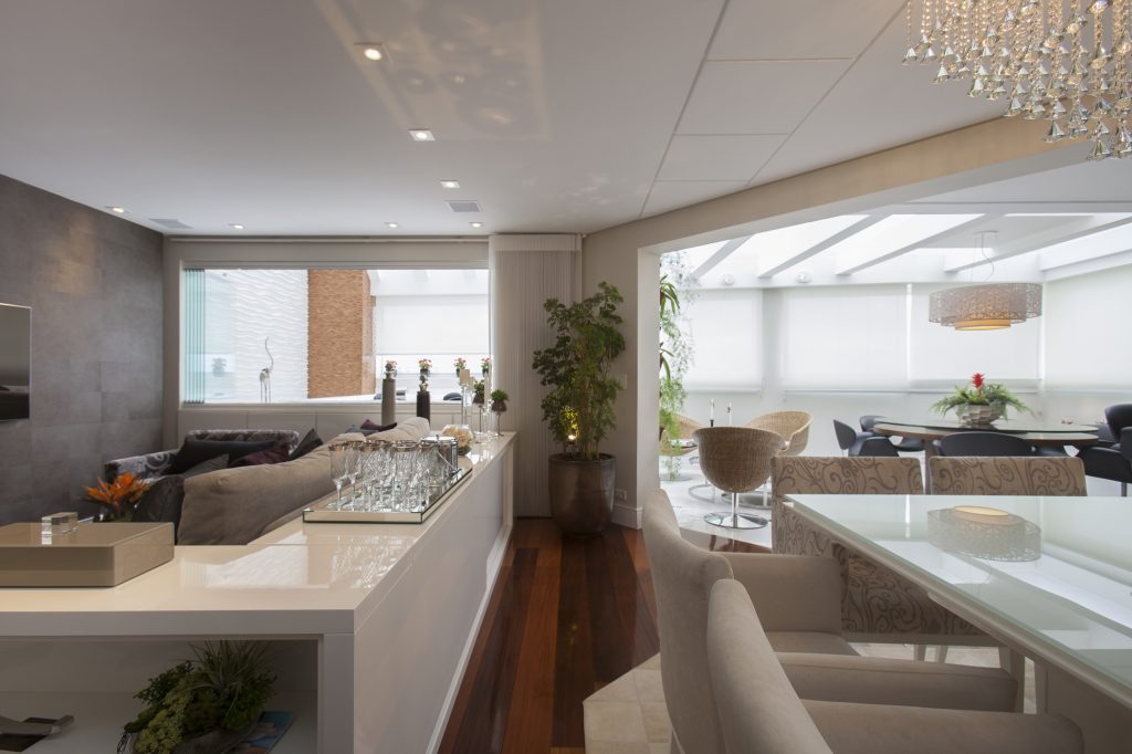 Deste ângulo é possível ver a integração total entre os ambientes internos e externos, com apenas o piso diferenciando os usos. Na sala, madeira quente e acolhedora. Fora, porcelanato durável e fácil de limpar. Entre o estar e o SPA, uma abertura na parede fechada por vidro faz a interação visual entre os espaços.